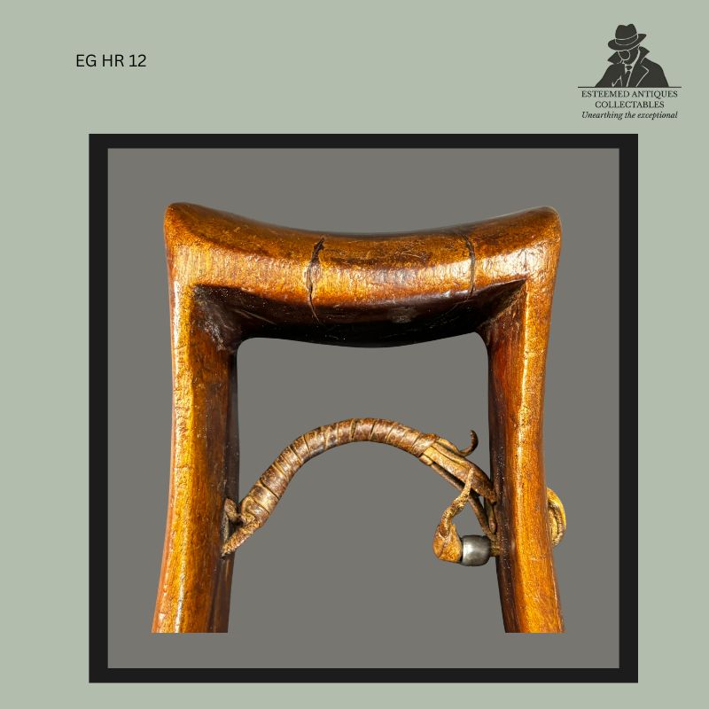 Detail view of an East African carved wooden headrest showing the original leather suspension element secured beneath the central support, with attached bead ornament and aged leather bindings, illustrating traditional construction, long-term use, and ownership history from the Egon Guenther Collection.