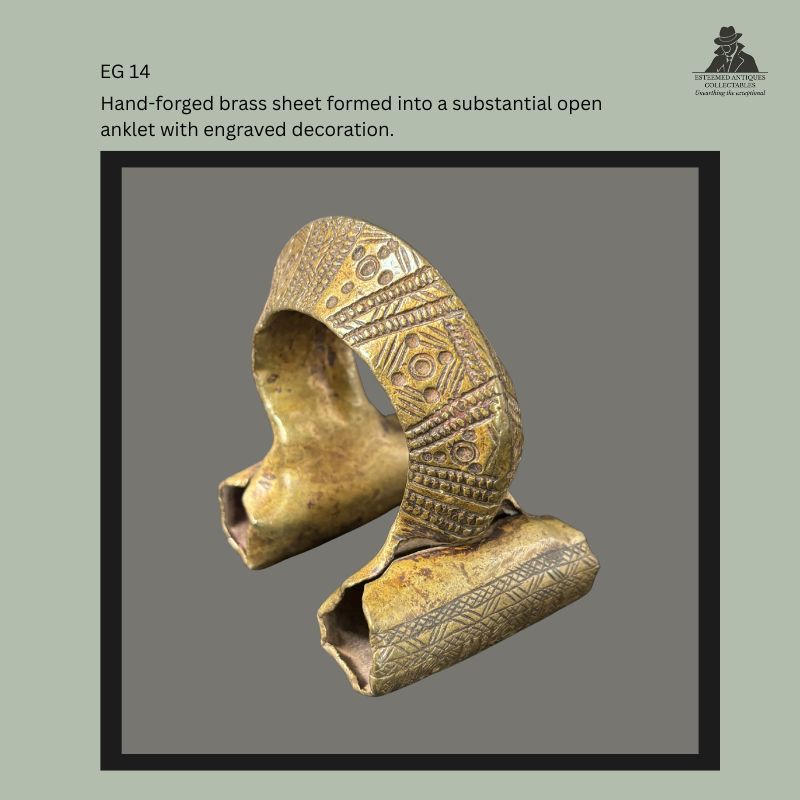Angled view of an Ethiopian brass sheet anklet with an open cuff form, hollow flared ends, and engraved geometric panels across the curved outer surface, from the Egon Guenther Collection.