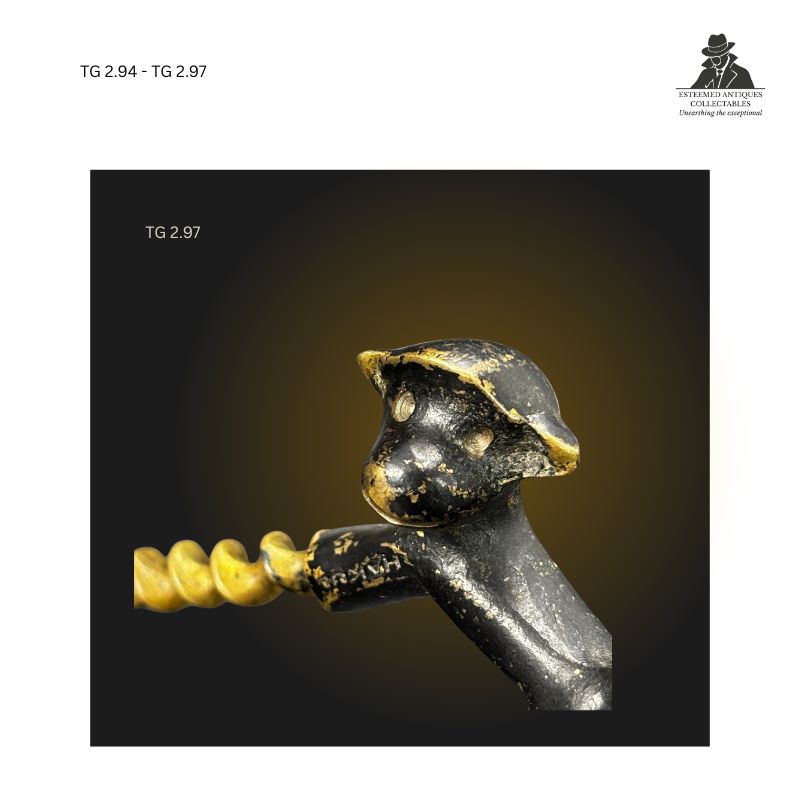 Front facial detail of a figural monkey corkscrew showing the cast metal monkey head forming the handle, with painted surface decoration and exposed spiral worm extending from the mouth area, viewed against a dark background.