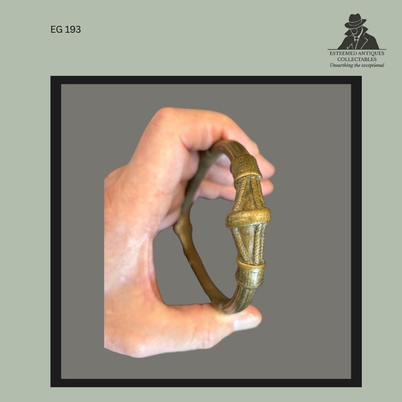 Cast brass or bronze anklet currency ornament attributed to the Frafra or Lobi peoples of West Africa, featuring tapered geometric terminals and ring-form construction, photographed in hand for scale, from the Egon Guenther Collection.