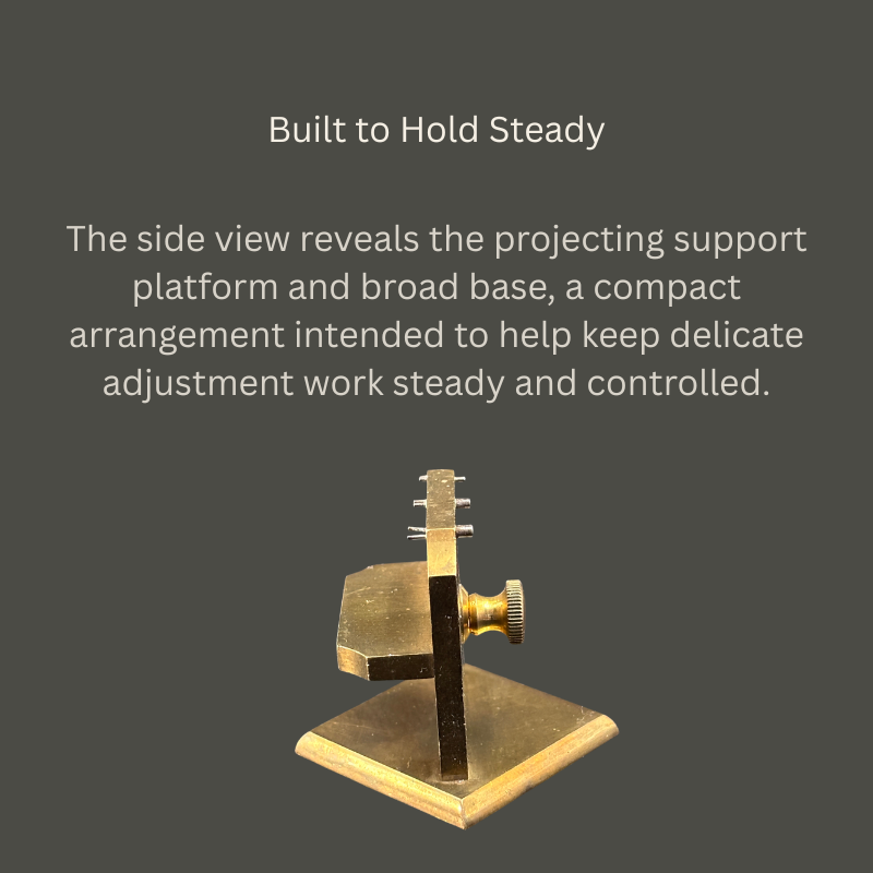 Side view of a vintage brass and steel watchmaker’s adjustment jig, showing its projecting support platform, upright plate, knurled adjustment screw and broad square base.
