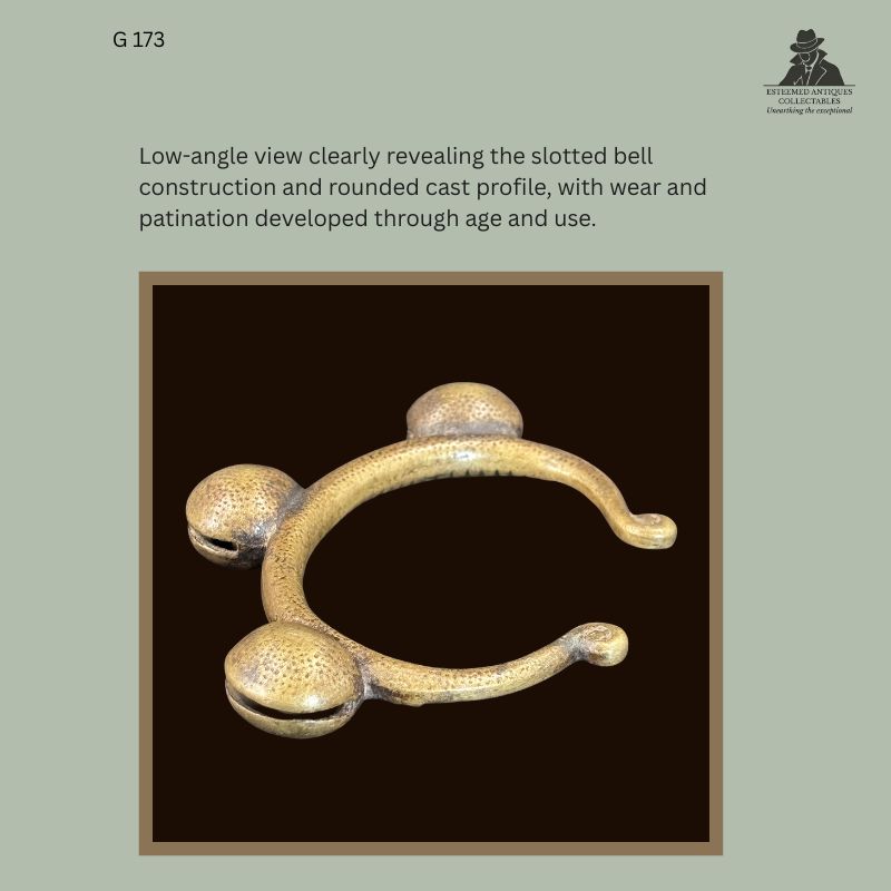 Low-angle detail of West African cast brass anklet EG 173 showing the hollow slotted bell forms, rounded cast profile, surface wear and age-related patination.