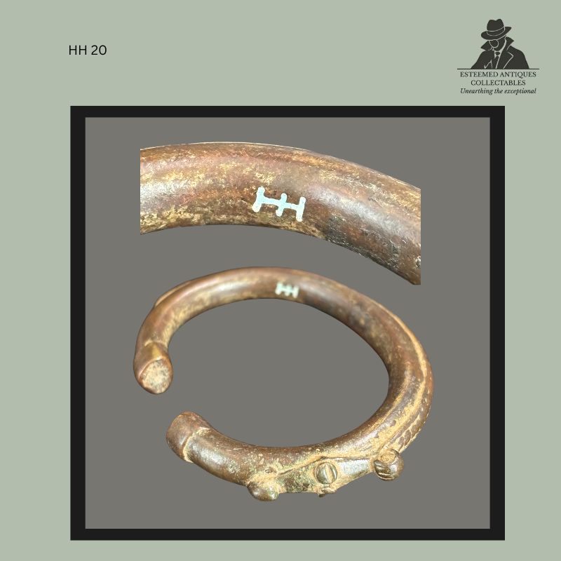 Cast brass open-form bracelet from West Africa, featuring a thick circular body with tapered terminals and small cast decorative projections near one end. The bracelet bears a painted HH inventory mark identifying former ownership within the Hans Himmelheber collection, and displays a dark aged patina consistent with long-term use and collection history.