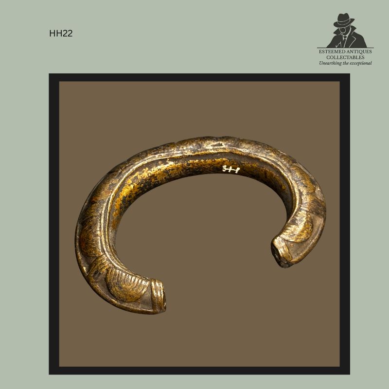 West African cast brass currency bracelet with open circular form, decorated with repeating crescent and geometric motifs along the outer surface, showing wear and patina consistent with traditional metalworking traditions, bearing a Hans Himmelheber collection inventory mark.