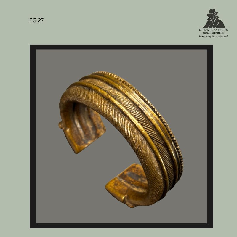 Profile view of a West African cast brass currency bracelet with open cuff form, displaying finely incised herringbone decoration, raised border bands, and worn patinated surface, from the Egon Guenther Collection by family descent to the Thomas Guenther Collection.