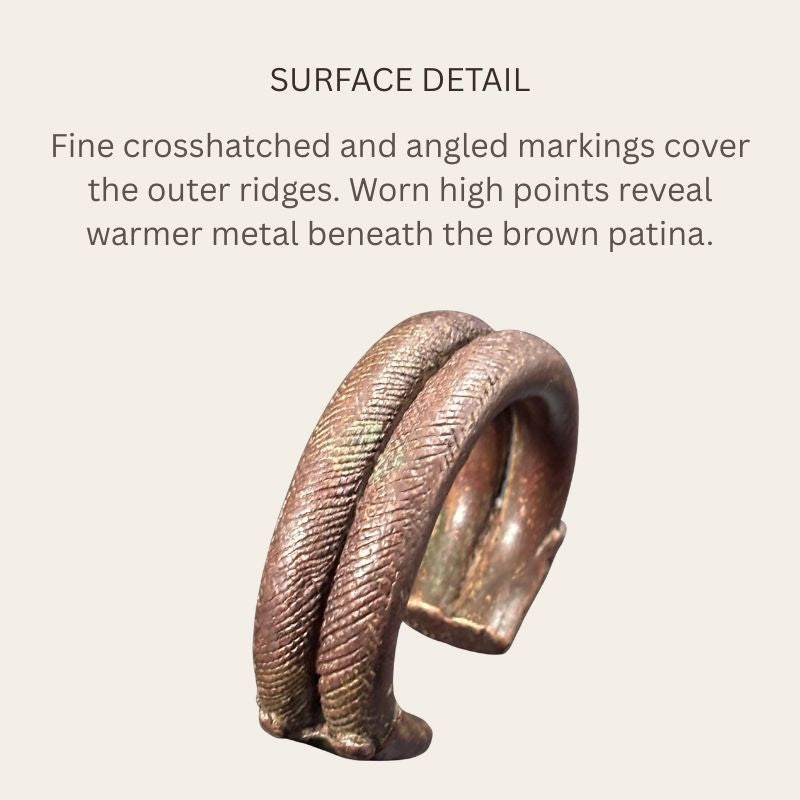 Close view of a West African copper-alloy penannular bracelet showing paired ridges with crosshatched and angled markings, brown patina, green oxidation and warmer exposed metal on worn high points.