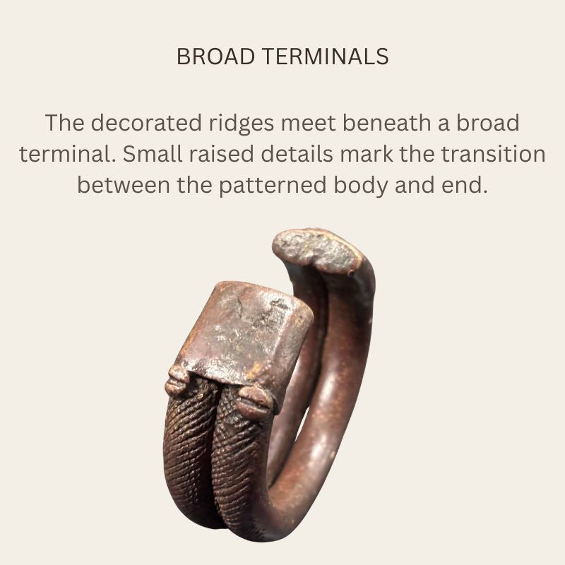 Oblique view of a West African copper-alloy penannular bracelet showing broad flattened terminals, paired patterned ridges, small raised details and a smooth inner surface.