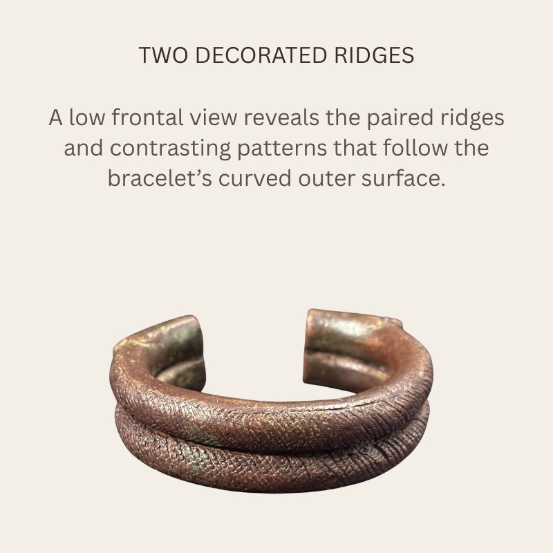 Low frontal view of a West African copper-alloy penannular bracelet, showing two rounded ridges with contrasting crosshatched and angled decoration, broad open terminals and brown patina.
